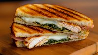 Grilled panini sandwich with turkey, cheese and pesto on wooden cutting board. Perfect for restaurant menus, food blogs and culinary presentations.