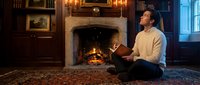 Young Caucasian man in cream sweater reading book by fireplace in cozy library room with wooden shelves, warm lighting creating peaceful evening atmosphere.