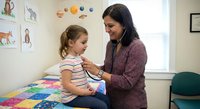 Hispanic female pediatrician examining young Caucasian girl patient with stethoscope in colorful medical office with educational posters and toys.