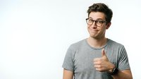 Young man with dark hair and glasses wearing gray t-shirt giving thumbs up gesture against white background for approval and success concepts.