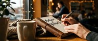 Person writing in gratitude journal with coffee cup during cozy holiday evening at home with warm lighting and festive decorations in background.