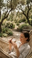 Woman relaxing in hammock holding sunglasses in shaded garden with blooming flowers and wooden bench, ideal for lifestyle and wellness content.