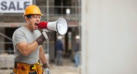 Construction worker with orange hard hat shouting through megaphone at building site. Male contractor using loudspeaker for safety announcement or team communication.