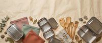 Flat lay arrangement of reusable food storage containers, wooden utensils, and kitchen accessories on neutral fabric background for sustainable living and eco-friendly home organization content.