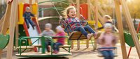Happy toddler girl with blonde curly hair enjoying swing at colorful playground during golden hour with motion blur of children playing in background.
