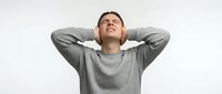 Young Caucasian man in gray sweatshirt stretching with hands behind head, eyes closed in relaxation pose against white background for wellness content.