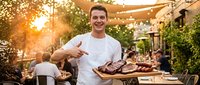 Young Caucasian male chef presenting grilled meat platter at outdoor restaurant terrace during golden hour sunset with string lights and dining guests.