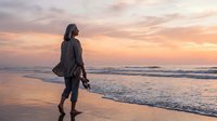 Young woman in wetsuit holding surfboard on beach at sunset, standing in shallow ocean waves during golden hour, ready for water sports and surfing adventure.