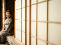 Mature Asian woman in traditional gray kimono sitting on wooden window sill, gazing thoughtfully through latticed shoji screen with warm natural light filtering through paper panels.