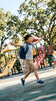 Happy Asian elementary school boy with backpack jumping with joy at colorful playground during sunny day back to school celebration outdoors.