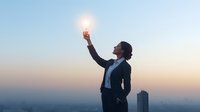 Young Asian businesswoman in suit holding glowing light bulb against urban sunset skyline, representing innovation, leadership and creative vision in business world.