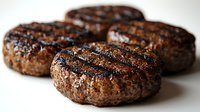 Grilled beef burger patties with charred grill marks and seasoning on light background, close up view showing juicy texture and caramelized surface.