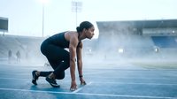 Athletic African American woman in starting position on blue track at stadium preparing for sprint race with dramatic lighting and smoke effects.
