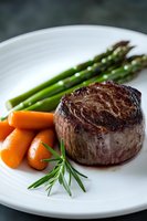 Grilled beef tenderloin steak served with fresh asparagus and baby carrots on white plate, garnished with rosemary sprig. Close up food photography.