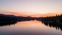 Serene lake at sunrise with mirror-like water reflecting golden and pink sky, surrounded by forested mountains and evergreen trees along shoreline for nature and landscape projects.