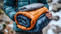 Person holding folded orange and blue down sleeping bags outdoors in winter camping environment, showing quality outdoor gear and equipment details.