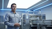 Professional Asian male technician in gray uniform standing confidently in modern industrial facility with advanced machinery and LED lighting.