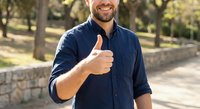 Happy Caucasian man in navy blue shirt giving thumbs up gesture outdoors in sunny park setting with natural bokeh background for positive lifestyle concepts.