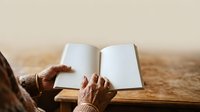 Senior person with aged hands holding open blank notebook on wooden table with neutral beige background, suitable for education, writing, journaling, memoir, and storytelling concepts.