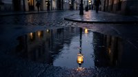 Wet cobblestone street at dusk reflects warm lantern lights and historic buildings, creating moody urban atmosphere for travel, architecture, and evening cityscape projects.