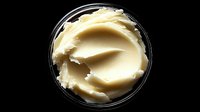 Creamy yellow shea butter in transparent glass container on black background, used in natural skincare and cosmetic formulations.