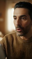 Middle-aged man with dark hair and beard wearing tan knit sweater, expressing surprise or concern with warm studio lighting backdrop for portrait and editorial use.