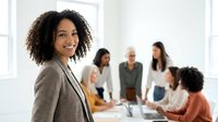 Confident African American businesswoman smiling in modern office with diverse team meeting in background for corporate presentations and leadership content.