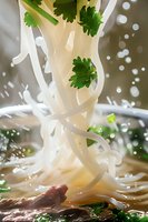 Fresh rice noodles falling into Asian soup with splashing broth, garnished with cilantro leaves, dramatic food photography perfect for restaurant menus and culinary advertising.