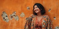Young Asian woman in colorful bohemian outfit smiling against vibrant orange textured wall background for lifestyle and fashion content.