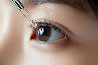 Close-up view of eye drops being administered with medical dropper, showing detailed macro shot of brown iris, eyelashes and smooth skin texture in soft lighting.