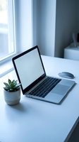 Modern laptop computer with blank screen on white desk next to small succulent plant in minimalist home office workspace with natural window light.