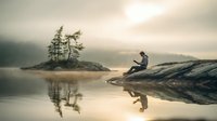 Young man sitting on rocky shore reading book at misty lake during sunrise, with small island and pine trees reflected in calm water.