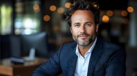 Confident middle-aged Caucasian businessman with beard smiling in modern office environment with warm bokeh lighting background for corporate presentations.