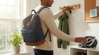 Young professional man with backpack organizing entryway, hanging jacket on wooden wall hooks in modern home interior with natural lighting.