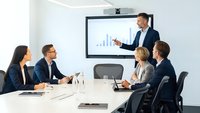 Professional businessman presenting financial data charts to diverse team during corporate board meeting in modern conference room.