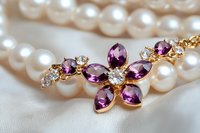 Elegant flower pendant with purple amethyst stones and diamonds on gold setting against pearl necklace background, macro jewelry photography on white silk.