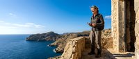 Senior Caucasian male hiker with backpack exploring ancient stone ruins overlooking dramatic blue Mediterranean coastline on sunny day.