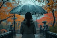 Asian woman with black umbrella standing on bridge in autumn rain. Moody atmospheric scene with orange fall foliage and lanterns creating mysterious ambiance.
