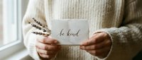 Person in cozy cream sweater holding a card with be kind calligraphy and dried lavender, soft natural light, mindfulness concept.