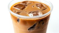 Refreshing iced coffee drink with milk in clear plastic cup, ice cubes floating on creamy brown surface, close up view against white background.