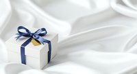 Small white gift box with navy blue ribbon bow and gold accent on luxurious white silk fabric background, suitable for holiday, wedding, and celebration design projects.