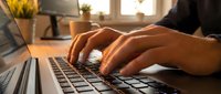 Close-up of male hands typing on a laptop keyboard at a wooden desk, warm sunlight in background, remote work concept.