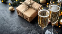 Festive celebration setup with sparkling champagne flutes, wrapped gift box and golden Christmas ornaments on dark stone background with bokeh lights.