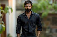 Serious South Asian man with beard wearing black shirt standing outdoors with blurred natural background.