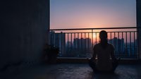 Silhouetted figure sitting on rooftop balcony overlooking sprawling cityscape at sunset, contemplative mood, urban solitude and reflection concept for lifestyle and wellness content.