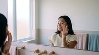 Young Asian woman applying skincare cream to face while looking in bathroom mirror during morning beauty routine for healthy glowing skin care.