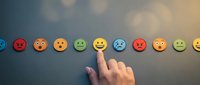 Hand pointing at happy yellow emoji face among colorful emotion icons representing customer satisfaction feedback and user experience rating system.
