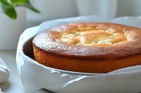 Freshly baked homemade cheesecake with golden brown top in white ceramic baking dish on light background, close up view of creamy dessert.