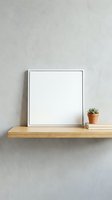 Blank white square frame mockup on wooden shelf with small succulent plant and books against textured gray wall for artwork display and home decor presentation.