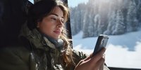 Young woman in winter coat using smartphone while traveling through snowy mountain landscape, sunlight illuminating forest scenery outside window.